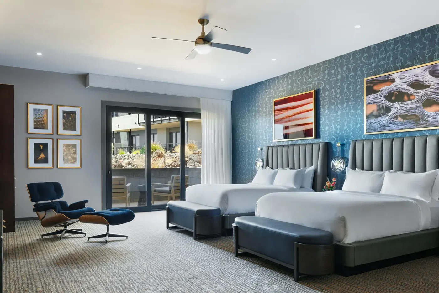 Representative bedroom with two queen beds, blue patterned wallpaper, a lounge chair and a sliding door to a patio